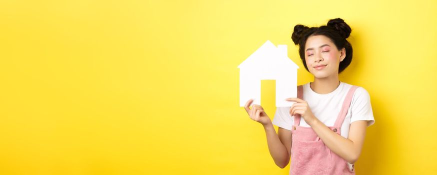 Real estate and family concept. Dreamy smiling asian woman with bright makeup, showing paper house cutout with closed eyes, daydreaming about buying property, yellow background.