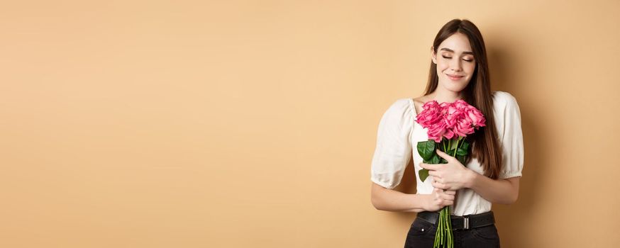 Valentines day. Tender and romantic caucasian woman close eyes dreamy, hugging bouquet of roses from secret admirer or lover, standing on beige background.