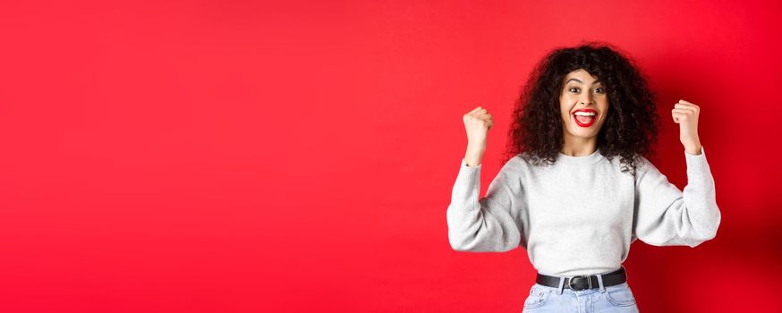 Cheerful lucky woman with curly hair, winning prize and scream yes with joy, raising hands up and celebrating, triumphing and cheering, standing on red background.