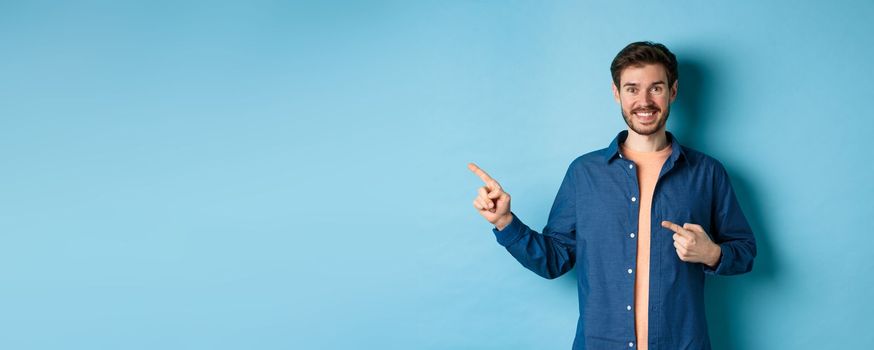 Happy smiling man pointing fingers left at logo, showing advertisement, standing on blue background.