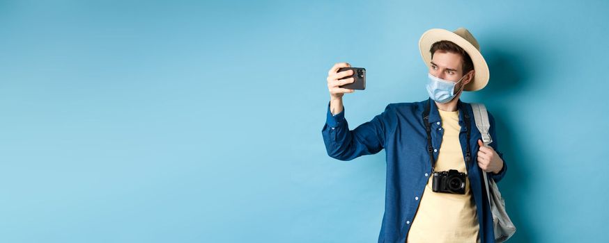 Covid-19, pandemic and travel concept. Male tourist in straw hat and medical mask recording video on smartphone during vacation, taking pics on summer holiday, blue background.