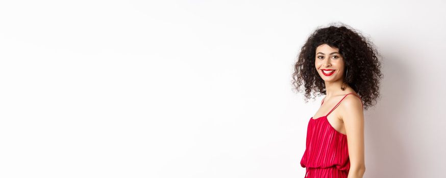 Profile of fashionable curly-haired woman in red dress, turn head at camera and smiling confident, standing over white background.