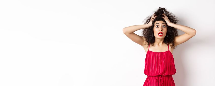 Shocked young woman holding hands on head and panicking, staring frustrated at camera, wearing red dress and makeup, white background.