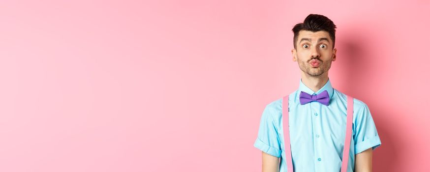 Funny young man waiting for kiss with puckered lips and silly face, standing on romantic pink background. Concept of love and Valentines day.