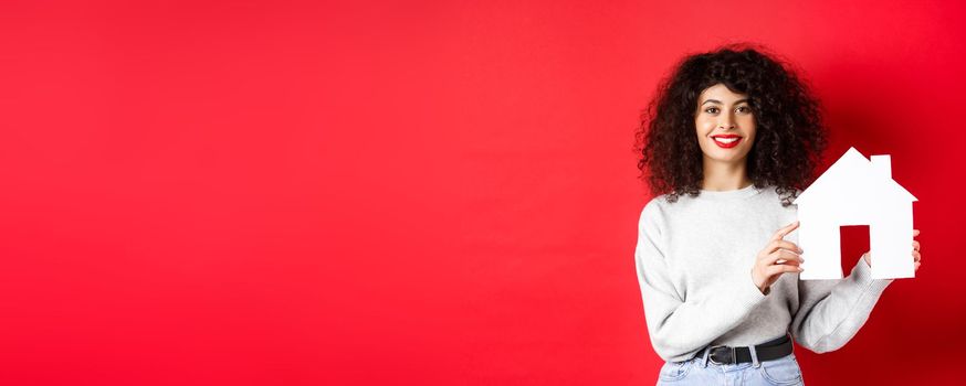 Real estate. Smiling caucasian woman with curly hair and red lips, showing paper house model, searching property, standing on red background.
