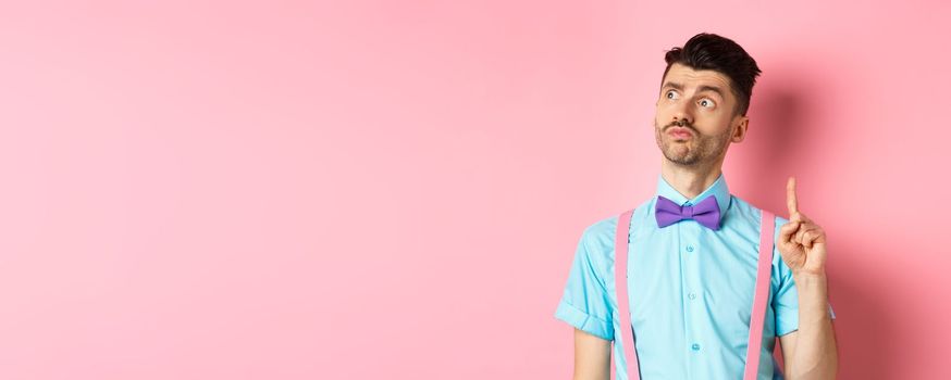 Pensive handsome man in bow-tie pitching an idea, raising index finger while looking away with thoughtful face, standing on pink background.