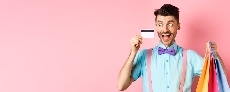 Cheerful funny guy looking at plastic credit card and smiling, buying gifts, holding shopping bags, standing on pink background.