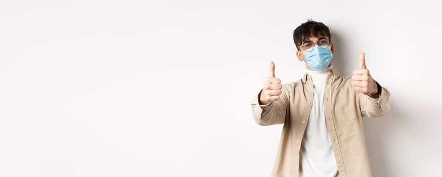 Coronavirus, health and real people concept. Smiling guy in medical mask showing thumbs up, wearing glasses, standing on white background.