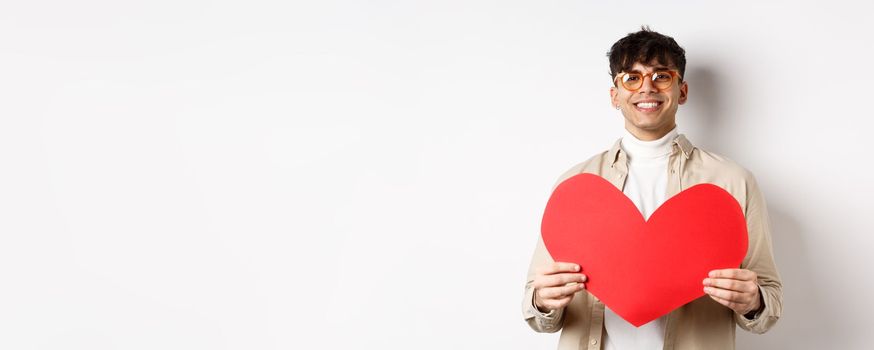 Attractive modern man smiling and looking at camera hopeful, holding big red Valentines heart, waiting for soulmate on lover day date, standing over white background.
