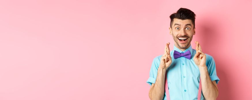 Happy guy making wish, holding fingers crossed for good luck and smiling at camera, waiting for good news, standing over pink background.