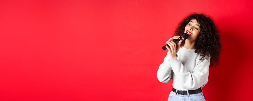 Leisure and hobbies concept. Stylish young woman singing karaoke, looking aside and holding microphone, performing song, standing on red background.
