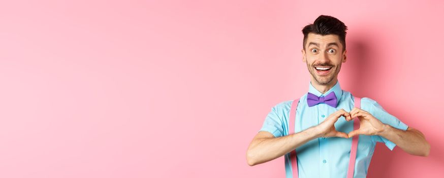 Passionate guy in funny bow tie saying I love you, showing heart gesture on Valentines day and smiling, expressing sympathy to lover, standing over pink background.