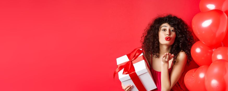 Romance and Valentines day concept. Pretty curly-haired girl in red dress sending her love, blowing air kiss at camera, holding gift from lover, standing near hearts on red background.