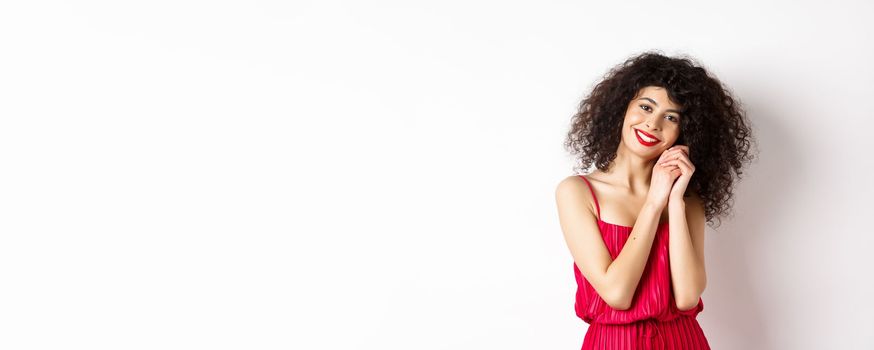 Lovely young woman with curly hairstyle, wearing red dress, looking at silly and tender thing, admire something, standing over white background.