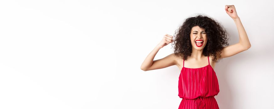 Cheerful emotive woman with curly hair, red dress, raising hands up and chanting, rooting for team, shouting wanting to win, standing on white background.