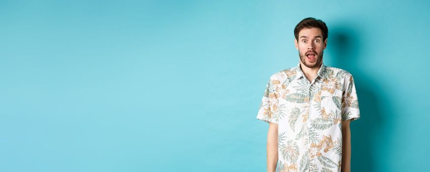 Summer holiday. Surprised guy in hawaiian shirt gasping amazed, checking out vacation offer, standing on blue background.