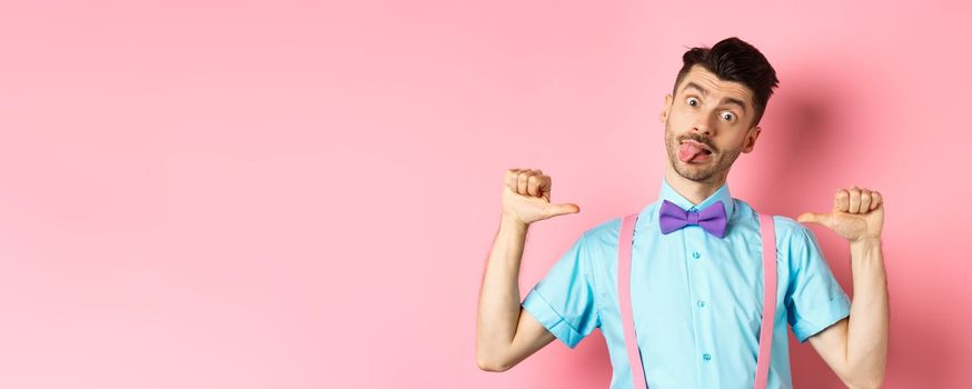 Funny guy in suspenders and bow-tie showing tongue, pointing at himself as if self-promoting, standing over pink background.