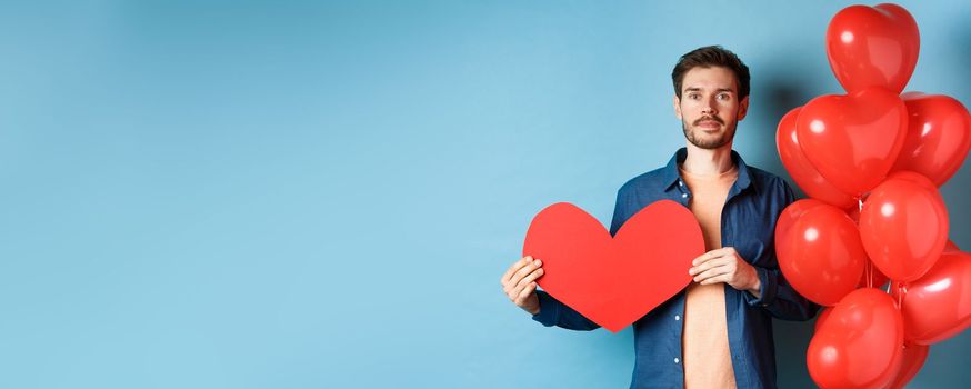 Valentines day and love concept. Young man showing red paper heart cutout and standing near romantic balloons, looking at lover, standing over blue background.