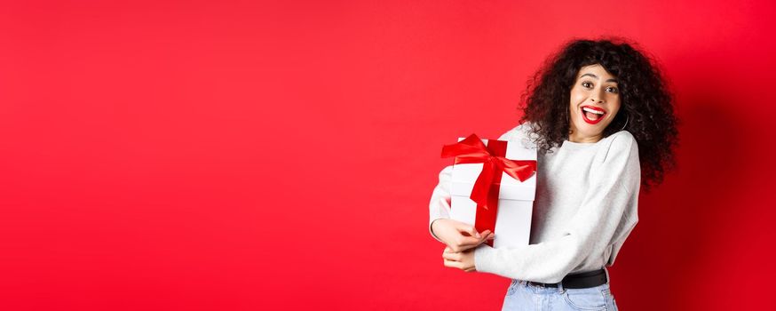 Valentines day. Happy girlfriend holding gift box from secret admirer, looking romantic at lover, receive present, standing on red background.