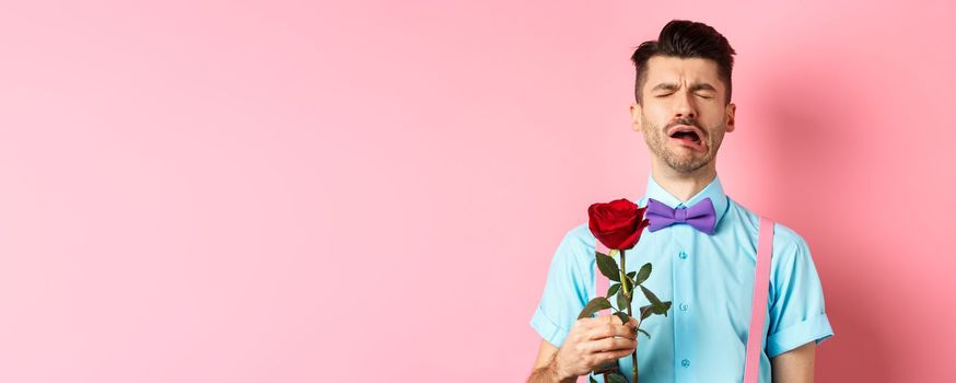 Heartbroken guy in funny bow-tie crying over girlfriend, standing alone with red rose on pink background and sobbing, break-up on Valentines day.