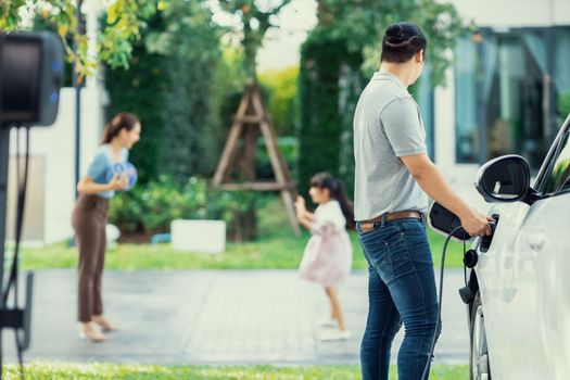 Focus image of progressive man charging electric car from home charging station with blur mother and daughter playing together in the background.