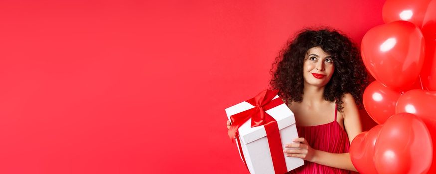 Valentines day and love concept. Beautiful woman with romantic makeup, holding gift box from secret admirer and looking at upper left corner dreamy, standing with heart balloons on red background.