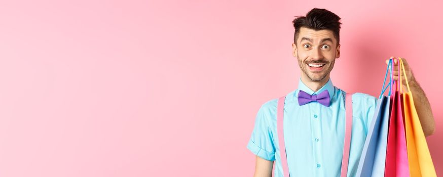 Image of happy guy on shopping, holding paper bags and smiling excited, shopper buying with discounts, standing on pink background.