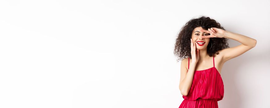 Beauty and fashion. Romantic woman with curly hair and red dress, showing v-sign and smiling happy at camera, standing on white background.
