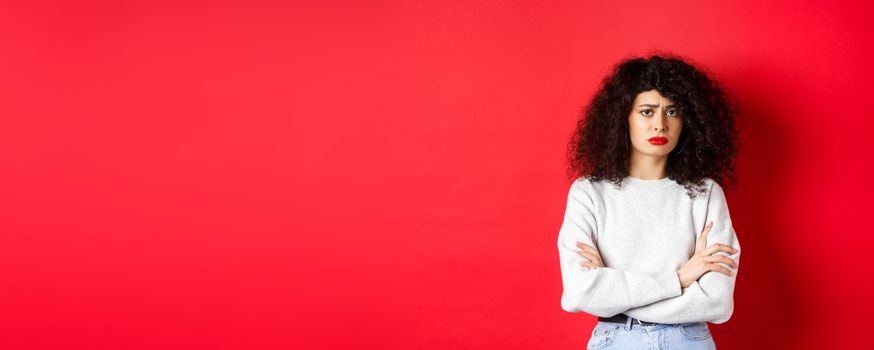 Sad and worried caucasian woman frowning, cross arms on chest and looking concerned, feeling bad, standing on red background.