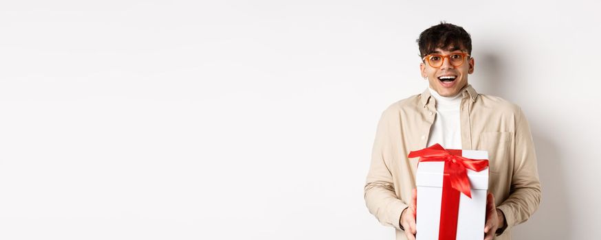 Surprised happy guy receiving a gift, holding present box and smiling amazed, standing on white background.