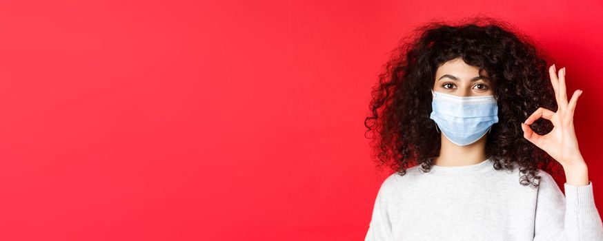 Covid and health concept. Confident young woman in medical mask showing OK gesture in approval, guarantee quality, standing on red background.