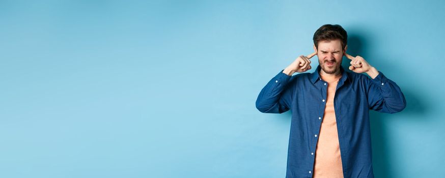 Annoyed young man block sound, shut ears with fingers and grimacing, disturbed by loud music, standing on blue background.