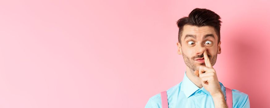 Image of funny caucasian guy picking nose and squint eyes, making dumb face, standing on pink background.