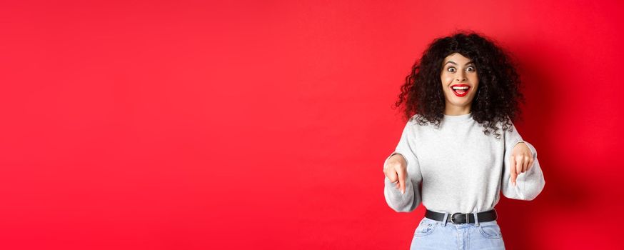 Excited woman pointing fingers down and smiling amazed, look here gesture, showing logo on red empty space.