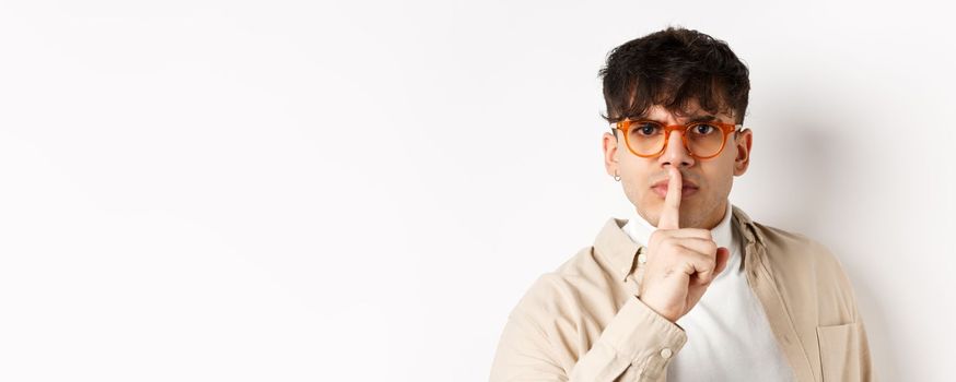 Close-up of grumpy young guy in glasses frowning and shushing, make taboo gesture with finger shuting lips, tell to be quiet, standing angry on white background.
