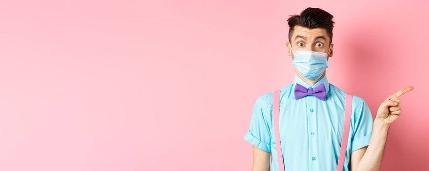 Coronavirus, healthcare and quarantine concept. Man looking surprised in medical mask, asking question and pointing right, curious about promo offer, standing on pink background.