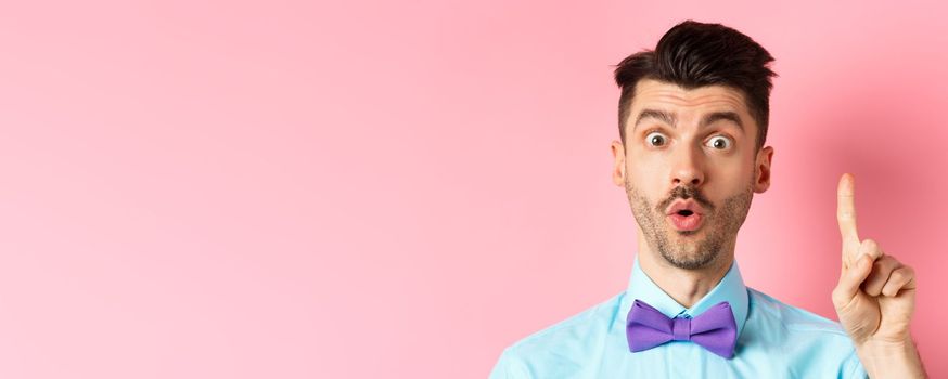 Close-up of excited funny guy with moustache and bow-tie, pitching an idea, raising finger to say suggestion, have plan, standing over pink background.