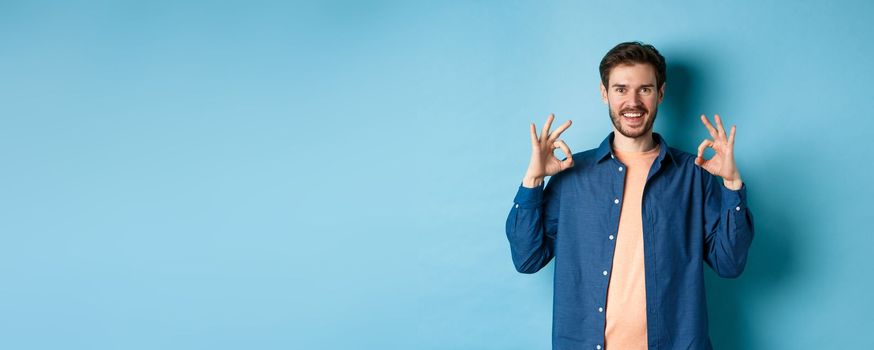 Satisfied young man smiling and showing okay gesture, approve something good, praising excellent thing, standing on blue background.