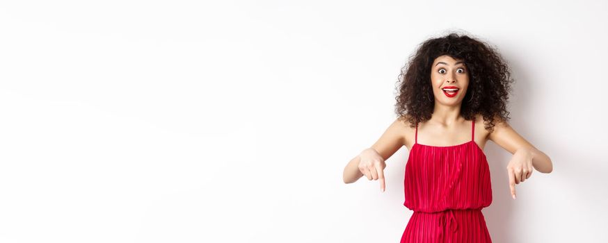 Excited woman with curly hair and red lips, wearing evening dress, gasping and pointing fingers down at super cool promo, standing on white background.