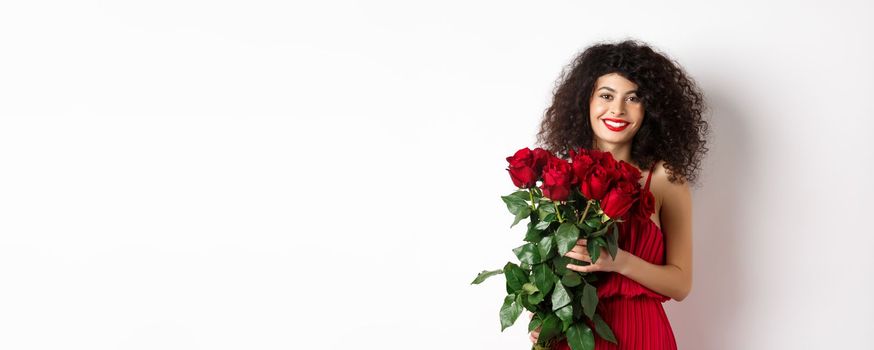 Gorgeous caucasian woman in red evening dress receiving bouquet of flowers. Girl with red roses smiling at camera, having romantic date on Valentines lover day, white background.