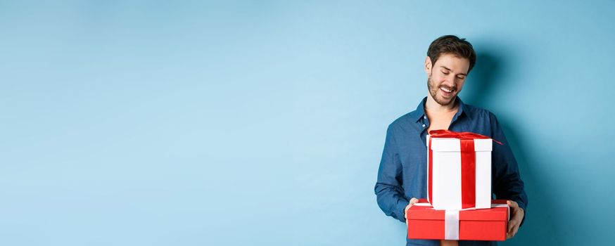 Romantic young man with beard, looking happy at gift boxes on valentines day, giving presents to lover, standing over blue background.