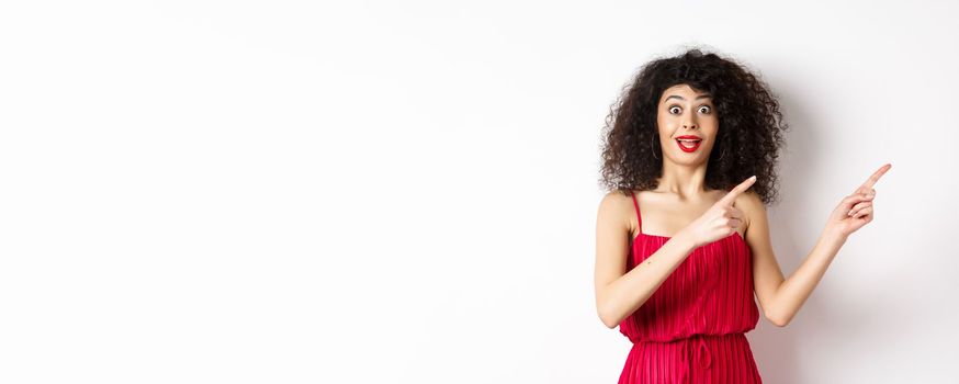 Surprised woman with curly hair, makeup and red dress, looking amazed and happy while pointing fingers right at logo, showing advertisement, white background.