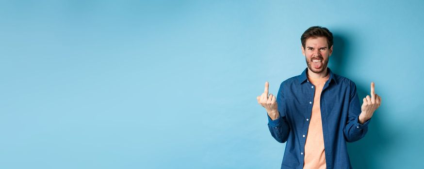 Ignorant and rude guy showing middle finger and smiling, say fuck you, standing on blue background. Copy space