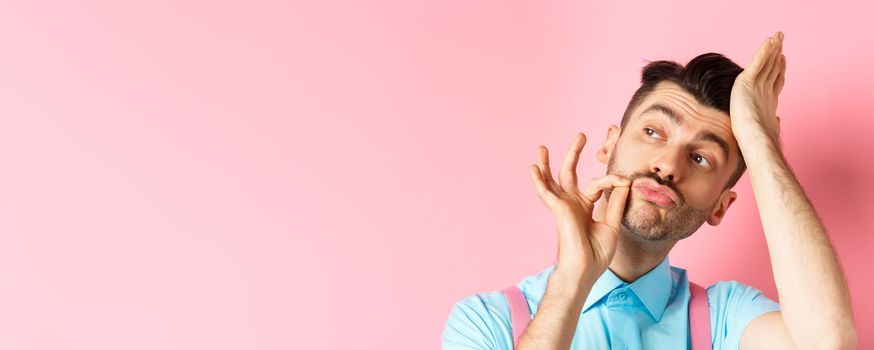 Close-up of handsome man fixing haircut and touching french moustache, looking aside confident, standing over pink background.