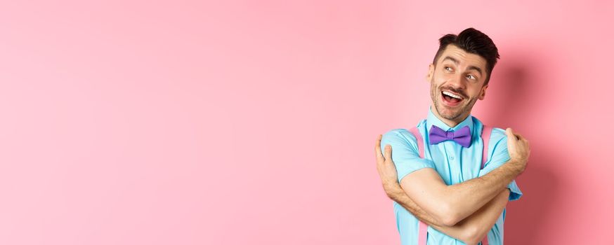 Happy and positive guy in bow-tie, hugging himself and looking left with cheerful smile, standing over pink background.