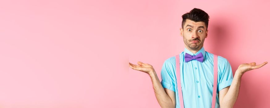 Image of confused guy in bow-tie and suspenders know nothing, shrugging shoulders and looking clueless, standing over pink background.