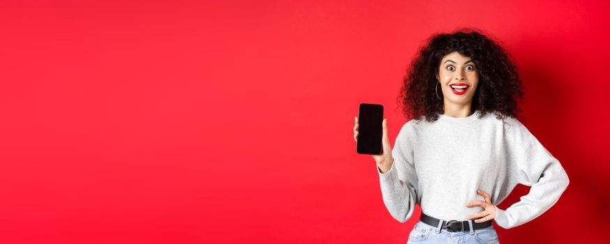 Excited woman with curly hair and red lips, showing empty smartphone screen and screaming from joy, standing on red background.