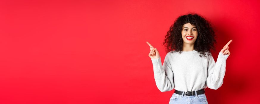 Attractive caucasian woman showing way, pointing fingers sideways at two promos, demonstrating variants and smiling, standing on red background.