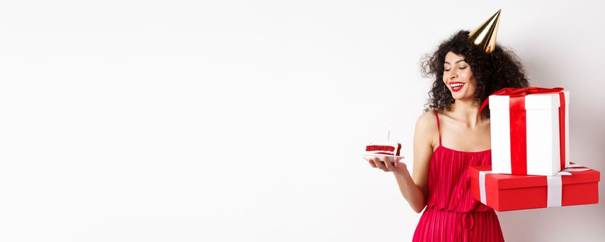 Cheerful smiling woman in red dress and party cone, looking happy at birthday cake, holding gifts, standing on white background. Copy space