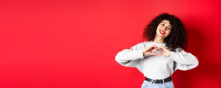 Beautiful young woman with curly hair showing heart gesture, say I love you and smile romantic at camera, standing on red background.